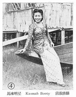 black-and-white image of Kasma Booty wearing a traditional outfit, leaning on a wooden boat, grinning directly at camera, with traditional stilt house in background