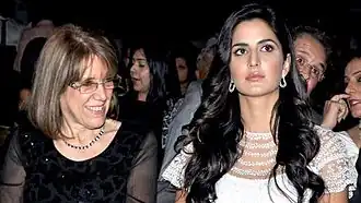 A woman is sitting next to an older woman in a crowded room. The younger woman is sitting on the right wearing a white dress, while the older woman is wearing a black dress.