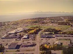 Aerial view of part of downtown Kenai. The intersection of Willow Street and Barnacle Way is in the center of the foreground. Cook Inlet and Mount Redoubt are in the background.