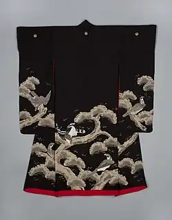 A black kimono with long sleeves viewed from the back. The kimono has three crests along the back and is lined in scarlet and has a thickly-padded hem. A painterly design of pine trees is seen across the bottom of the sleeves and from the waist down. Hawks are depicted as perching on the thick branches.