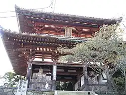 A two-storied wooden gate with white walls and faded red colored beams. There are two guardian statues in the side bays of the lower floor.