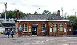 A small red-brick station building as viewed from some distance away across a car park. The building has a closed black double door with a sign above reading "Knebworth", as well as a phone, post-box, and defibrillator. The railway lies behind the building, which is clearly not in use as a station building operationally.