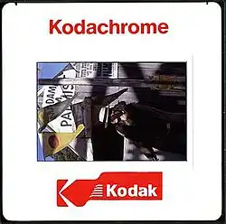 "A square white plastic frame, bearing the red text "Kodachrome" and a red logo bearing the word "Kodak", surrounds a portrait (rotated 90 degrees counter-clockwise) of a young woman wearing a white hat. She stands in front of a wooden building. Two triangular flags hang to the left, and the text "Madam M Palmist" is visible in the centre-top."
