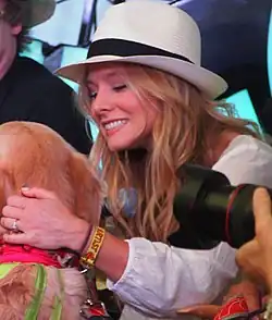 Photo of Kristen Bell wearing a light hat fedora hat, resting her left hand on the neck of a light brown dog
