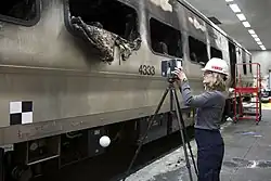 A woman wearing a white helmet on the right sets up a device on a tripod, pointing it toward a railcar on the left with "4333" on its side