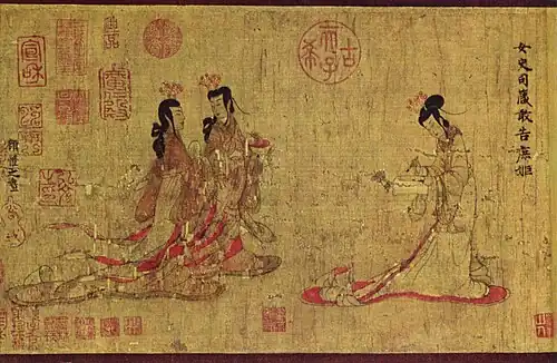 Two ladies walking towards another lady standing at a writing table with a writing brush in her hand