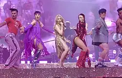 Minogue singing on stage in a golden dress surrounded by dancers in colorful outfits