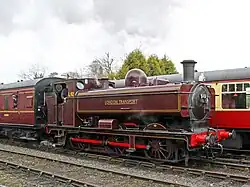 A black pannier tank locomotive is travelling from right to left, bunker first, along a single track on the side of a wooded hill. The locomotive is pulling five passenger carriages painted cream and brown, and a plume of smoke is passing over the first two carriages. The image of the train is reflected the river in front of the train.