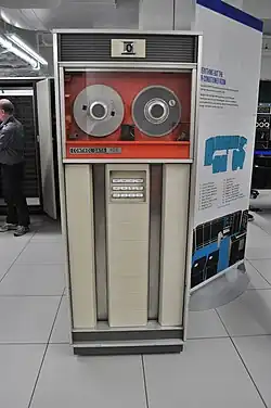 A large cabinet, about the size of an upright refrigerator, with a glass-covered top part holding two reels of magnetic tape, and a bottom part with control buttons framed by vertical channels.
