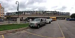An elevated train station crosses a busy road in the background; in the foreground, a silver SUV is parked.