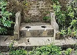 A stone bench-like construction with two holes in it, in front of a brick wall. There are plants growing around it.