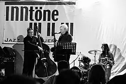 A monochrome image trio of musicians (tenor saxophone, drums and upright bass) performing.