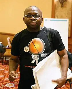 Leo Igwe wearing a black print t-shirt, carrying a bundle of papers under one harm, with a bag on one shoulder, lips pursed, looking right of camera
