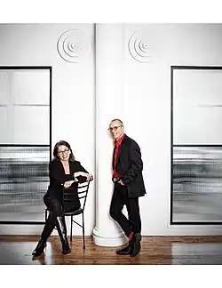 Seated woman leaning on a chair back next to a man leaning against a wall