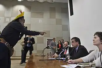 A man wearing traditional garments is standing, speaking and pointing directly at the three people sitting behind a table in front of him.