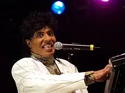 An African American man singing into a microphone, the microphone is being held up by a stand. He is wearing a cream color suit that has black pin stripes.