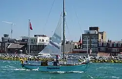 blue-hulled two-masted yacht with mainsail partly set passing a rocky shoreline with buildings beyond