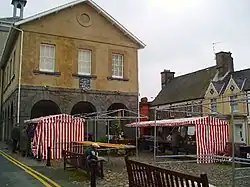 A couple of market stalls with red and white striped awnings in front of a stone building with three arched windows above stone arches