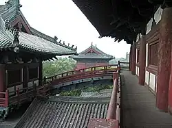 Several wooden buildings with grey tile roofs, connected by an arched bridge. A forest can be seen behind the buildings.