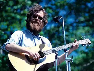 Loudon Wainwright III wearing sunglasses and playing an acoustic guitar on on outdoor stage