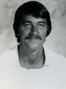 A young white man with a mustache smiling at the camera while wearing a white shirt.