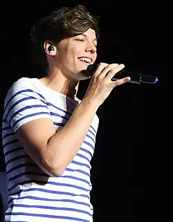 A young adult male wearing a white shirt with horizontal blue stripes, smiling and singing into a microphone