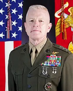 Official 2024 portrait of LtGen Paul J Rock, Jr, Director, Marine Corp Staff. Photograph of a Caucasian male with white hair in high-and-tight cut, wearing his Marine Corps olive green and khaki service uniform, with three silver stars on his shoulder epaulettes and a full left chest of military ribbons and medals. A US Flag is on his right and a Marine Corps flag on his left.
