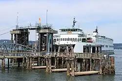 A ferry boat stopped at a wooden pier with a bridge, pilings, and a tower.