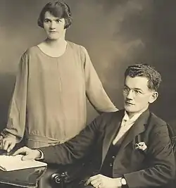 "Greyscale photograph showing a woman in a dress and with a fashionable hairstyle; she is standing to the right of and slightly behind a bespectacled man wearing a suit and breasted jackey seated on a chair with his right hand touching a white book on a table; each person is looking a little to their right