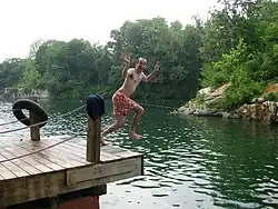 A man in a swimsuit jumps off a dock mid-air laughing into Beaver Dam.