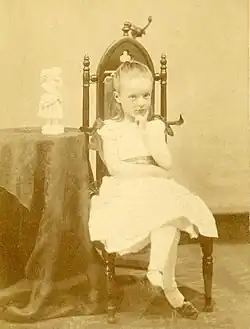 A small child in a white dress posed in a chair. She is looking into the camera and resting her chin in one hand. Next to the chair is a table on which rests a small figurine.