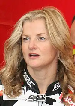 Tall slim woman with long blonde hair wearing multi coloured racing leathers standing next to an historic BMW motorcycle in the company of an older man