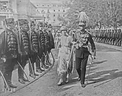 A woman in full dress and a man in army uniform walk down a street surrounded by soldiers.