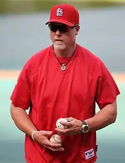 A man in a red athletic shirt holds a baseball in each hand. He is wearing a red cap and sunglasses.