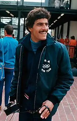 Mark Spitz turning slightly to the side and smiling while holding a camcorder in one hand. Shown from about the waist up.