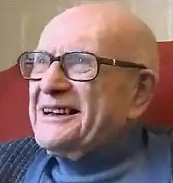headshot of white caucasian male, approx. 80-85 years old, bald, smiling, wearing thick spectacles and a blue sweater