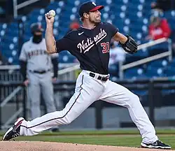 A man in white pants and a navy blue baseball jersey with "Nationals" written across the front throws a pitch.