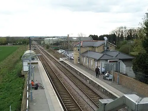 A view overlooking a station as taken from the footbridge over it on the left-hand side. There are two platforms, one either side of two tracks. On the right-hand platform there is a yellow-brick station building that is a mix of one and two storeys. Overhead line equipment, signals, and lamppostts are also visible.