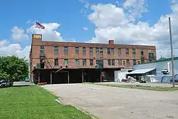 The same brick building seen from a different angle. An American flag flies from a short tower at the left end, and a flat roof projects over part of a parking area on that side at ground level. A one-story white building is at the right.