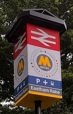 A station sign with a large British Rail logo, a large M with the text 'Metro' underneath, a Park and Ride indicator and the name 'Eastham Rake' underneath.