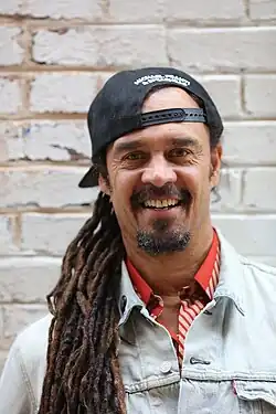Michael Franti wearing a light denim jacket over an orange shirt smiling for a portrait against a white brick wall