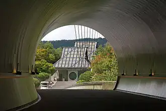 A passageway at the Miho Museum. A curving circular tunnel opens to reveal a building with a tall sloping roof and a circular window in the front door.