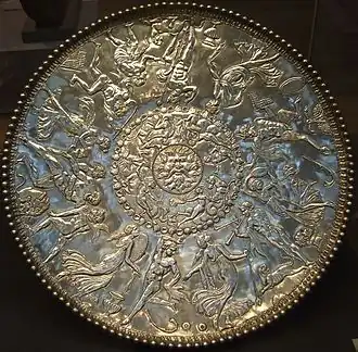 A large silver dish, decorated in bas relief in three rings. In the center ring, whose diameter is roughly 1/7 of the whole dish, is the face of a full-bearded man, with four bird heads pointing outwards along the diagonals. Around the second ring, frolicking sea creatures such as nereids are depicted; this ring's diameter is roughly 3/7 of the dish. Around the outermost ring, humans and a faun are depicted as dancing, making music, drinking and enjoying other pastimes.