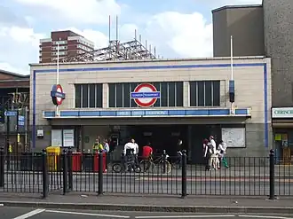 A grey-bricked building with a rectangular, dark blue sign reading "MILE END STATION" in white letters all under a light blue sky with white clouds