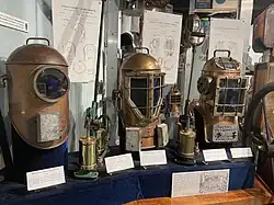 Reproduction of the 1715 John Lethbridge "barrel" design of diving equipment with educational material, photographs and drawings on wall in background