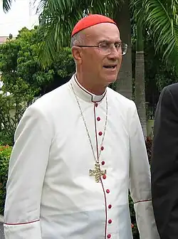 A white man wearing a white cassock and a red cap.