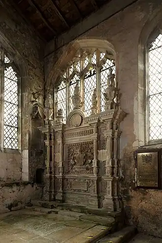 This image shows a deteriorating Renaissance wall monument of c.1570 to the Griffin family at Braybrooke, Northamptonshire, situated within a medieval church chancel with diamond-paned windows. The elaborate stone monument features classical architectural elements including paired columns, decorative urns, and heraldic shields, but shows visible signs of weathering and damp damage. Moss and discolouration are evident on the stonework, whilst the monument's base shows staining from moisture ingress. The surrounding church fabric also displays deterioration, with damaged wall surfaces and evidence of water penetration. This monument exemplifies the conservation challenges facing England's ecclesiastical heritage, where declining resources and building maintenance issues threaten the preservation of irreplaceable sculptural artworks. The contrast between the monument's original grandeur and its current condition illustrates the urgent need for conservation intervention to protect such culturally significant works for future generations.