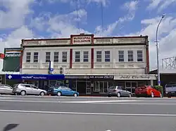 A photo of a two-storey building with white paneling and red highlights taken from across a street. A sign at the top reads Municipal Building. Signs for various stores line the first floor of the building