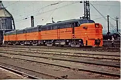 Three ALCO FA locomotives parked on a track in the yard. A railroad worker is standing near the lead locomotive.