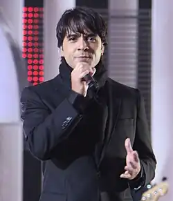 A man staring ahead with semi-long hair, wearing a black coat and a black scarf, holding a microphone in his left hand.